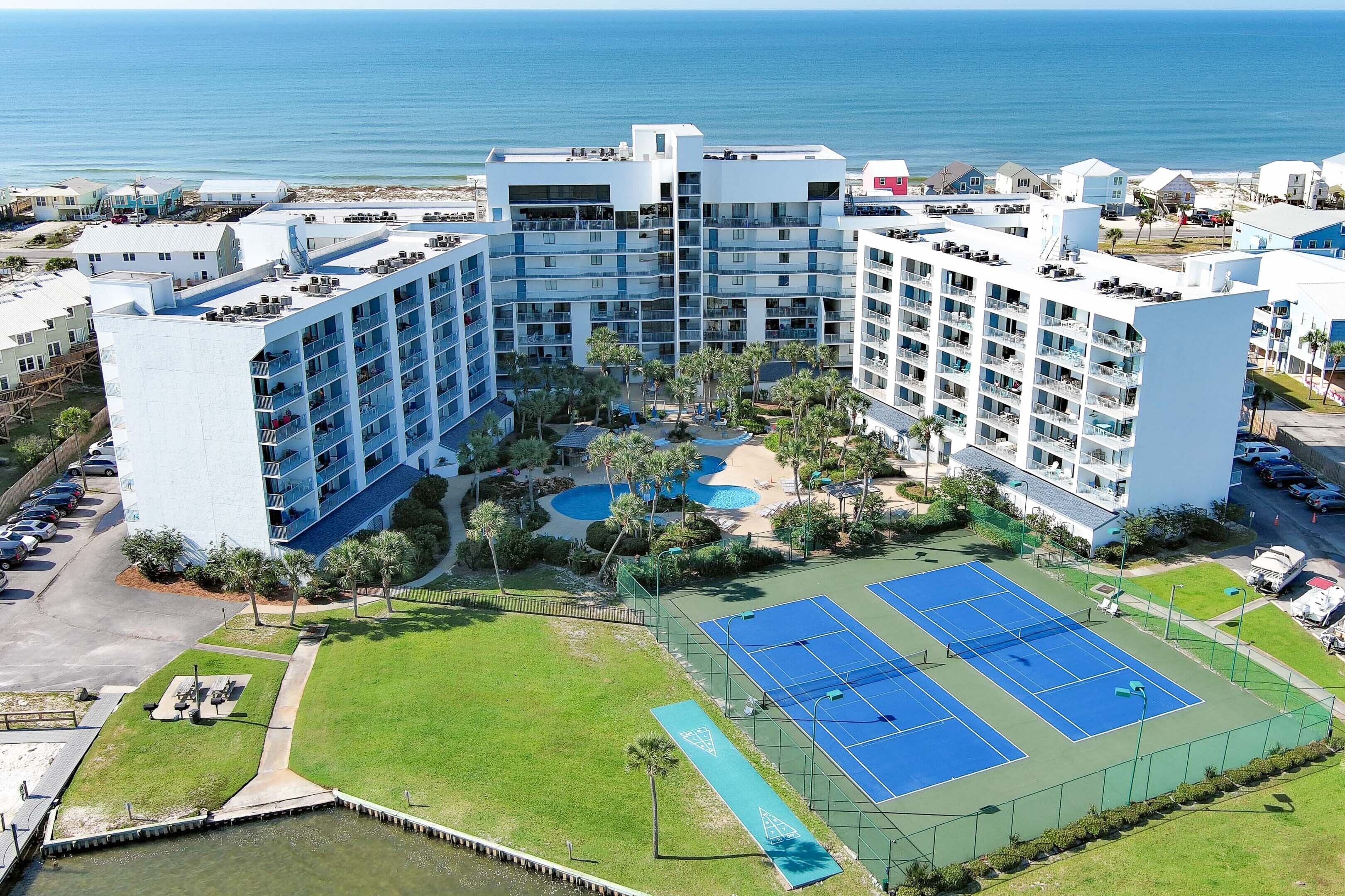 A large hotel with multiple floors, a swimming pool, tennis courts, and a view of the ocean.