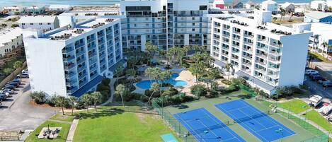 A large hotel with multiple floors, a swimming pool, tennis courts, and a view of the ocean.