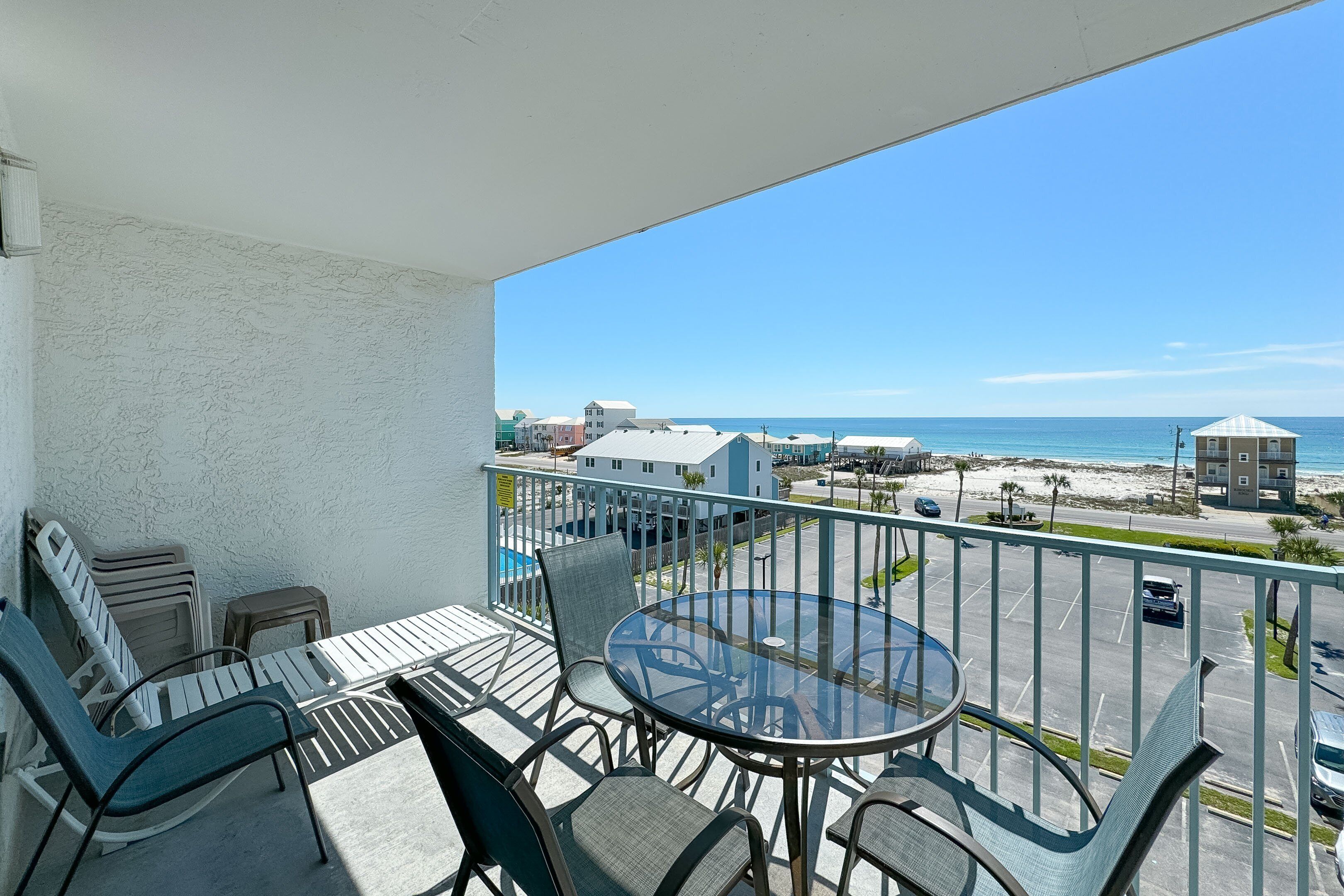 A balcony with a glass table and chairs, overlooking a beach and parking area.