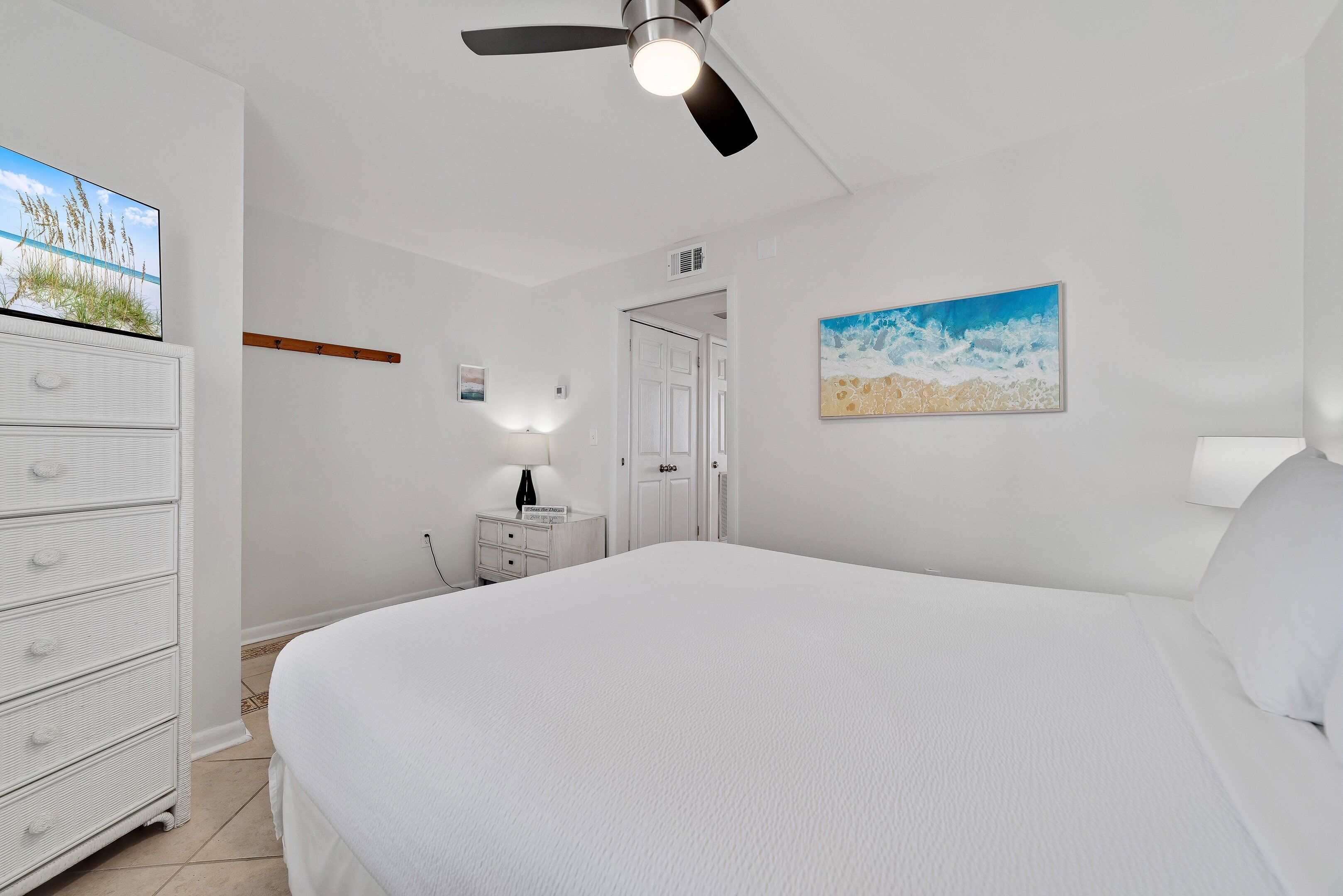 A bedroom with a large bed, a ceiling fan, a dresser, a nightstand, and a painting on the wall.