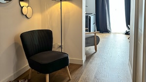 Une chambre d’hôtel moderne avec un fauteuil noir, une lampe sur pied, un miroir rond et une vue sur le couloir et la salle de bain, le tout visible à travers une fenêtre ornée de rideaux foncés.