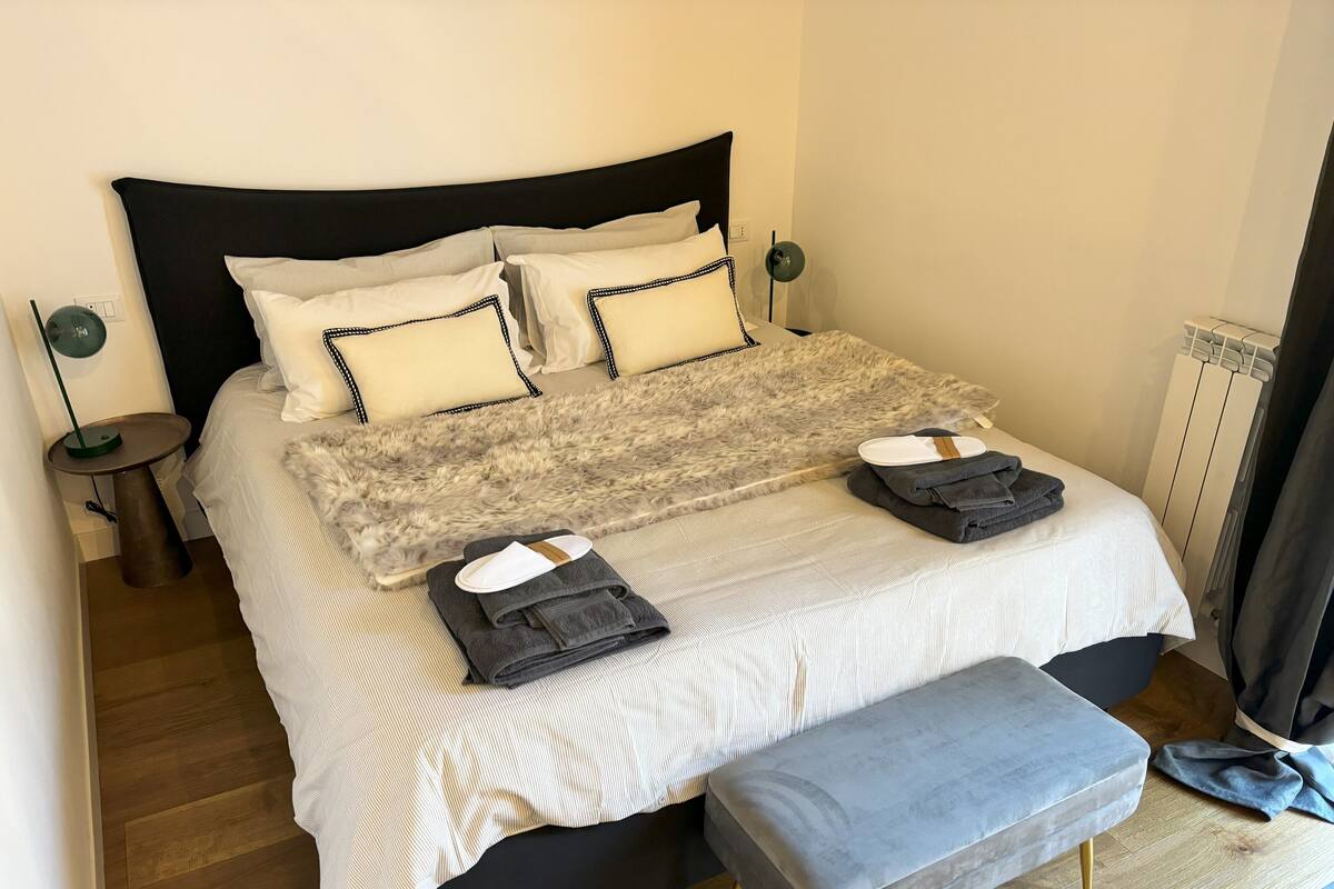 A neatly made bed with a fluffy blanket, two pillows, and folded towels on a grey upholstered bench.