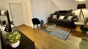 Un salon avec un canapé gris foncé, une table basse ronde, une chaise noire et un lampadaire. On y trouve une porte blanche et une petite plante posée sur une table.