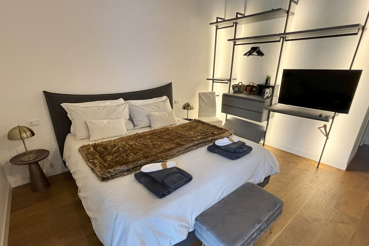 A modern bedroom with a large bed, a flat-screen TV, a desk, and a chair.
