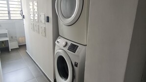 Uma lavanderia com máquinas de lavar e secar empilhadas, piso revestido e janela com persianas.