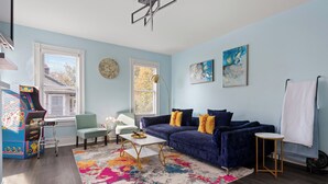 Ein Wohnzimmer mit blauer Wand, einem blauen Sofa, einem bunten Teppich, einem Couchtisch und einer Spielkonsole.