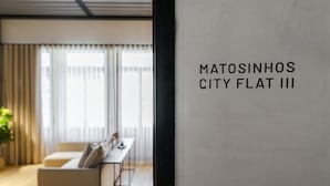 A modern interior with a sofa, a table, and a window with curtains. Text on the wall reads 'Matosinhos City Flat III'. - Feel Porto Matosinhos City Flat III (Matosinhos)