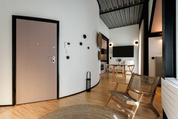 A modern interior with a curved wall, a wooden floor, a seating area with a woven chair, a small table, and a flat-screen TV mounted on the wall. - Feel Porto Matosinhos City Flat V (Matosinhos)
