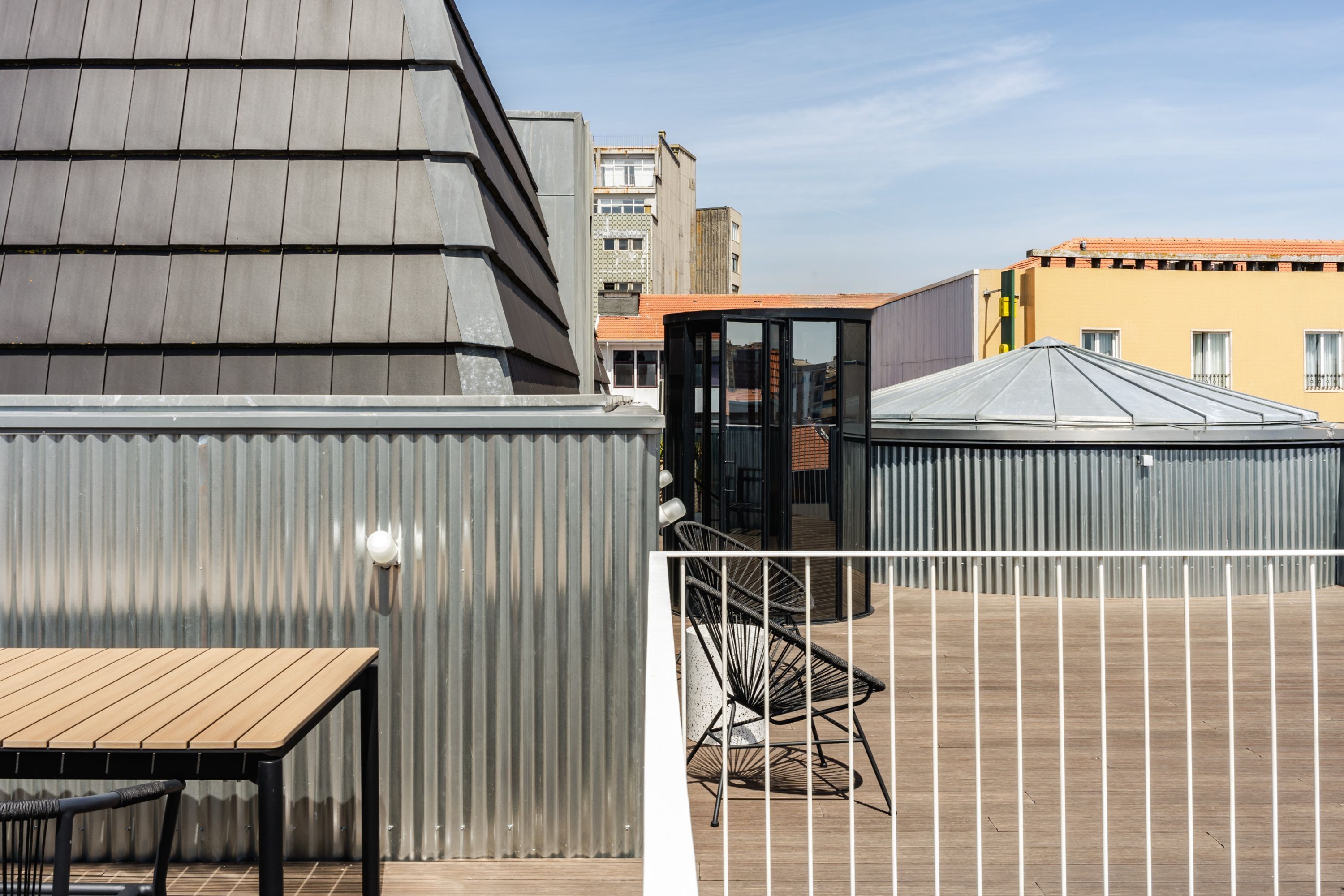 Una terraza en la azotea con una mesa y sillas de madera, una cerca metálica y edificios al fondo.
