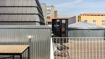 Una terraza en la azotea con una mesa y sillas de madera, una cerca metálica y edificios al fondo.