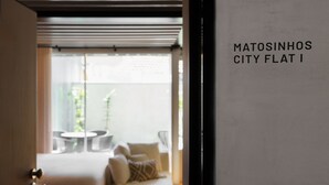 Une pièce avec un canapé et une petite table, une fenêtre avec des stores, et une pancarte indiquant « Appartement Matosinhos City I ».