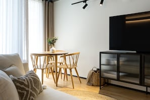 Een moderne woonkamer met een bank, een eettafel met stoelen, een televisie op een kast, en een plant op de tafel.