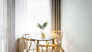 Une table ronde en bois, accompagnée de quatre chaises, un vase blanc orné d’une plante et des rideaux légers.