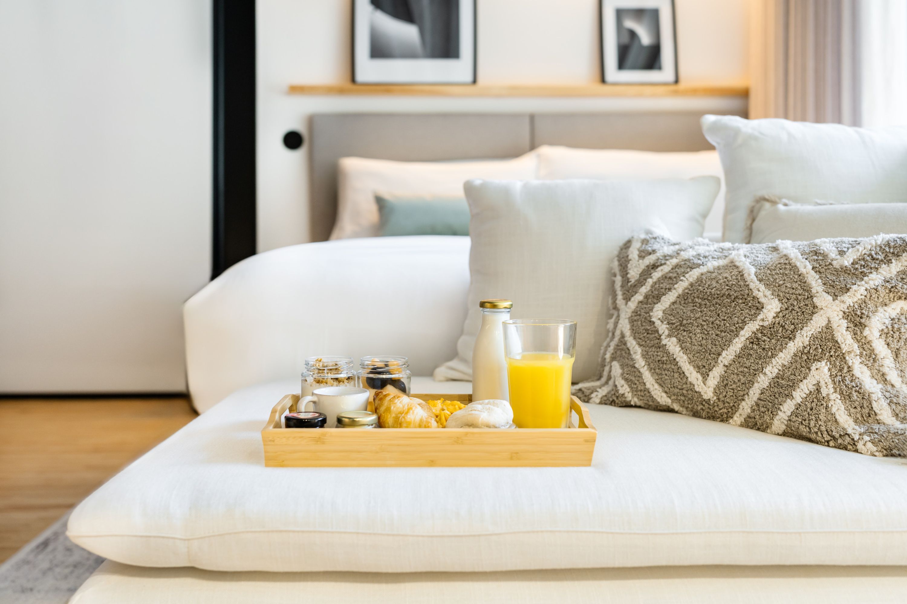 A neatly arranged breakfast tray on a sofa with a bed in the background, a bedside table, and a wall-mounted shelf with pictures.