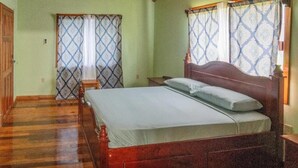 A bedroom with a wooden bed, a nightstand, a wooden wardrobe, and a window with patterned curtains. - Condo Caribbean Dreams (Dangriga)