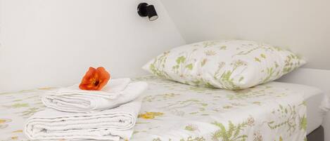 A neatly made bed with a floral-patterned bedspread, a folded white towel, and a single orange flower on top. There is a black wall-mounted light fixture above the bed.