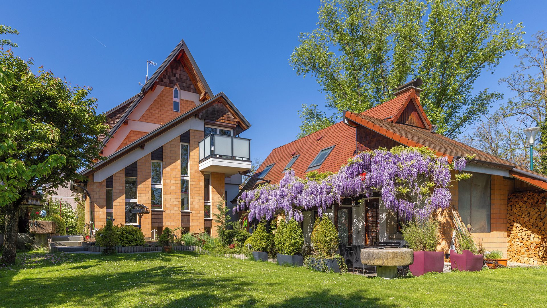 Une maison à deux étages, avec un toit recouvert de tuiles rouges, un balcon et un jardin orné de glycines aux fleurs violettes.