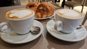Duas chávenas de café com arte de latte em formato de coração sobre pires brancos, acompanhadas por croissants num prato.