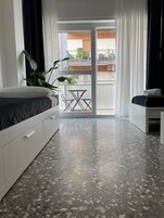 Una camera da letto con un letto, un tavolino, una sedia, una pianta in vaso e un balcone con vista sugli edifici.