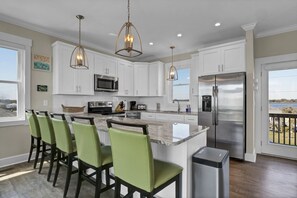 A modern kitchen with white cabinets, a stainless steel refrigerator, a large island with bar stools, and pendant lighting.