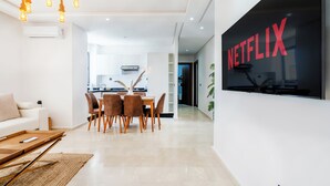 A modern living room with a dining area, a kitchen in the background, and a large TV screen displaying the Netflix logo. - Cozy Studio Near Train Stations & Hassan II Mosque (Casablanca)