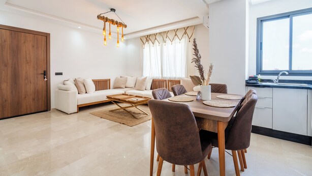 A modern living room with a sofa, dining table, and chairs. There is a wooden door, a window with curtains, and a hanging light fixture. - Cozy Studio Near Train Stations & Hassan II Mosque (Casablanca)