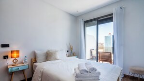 A hotel room with a bed, a nightstand with a lamp, a water bottle, and a view of a building through the window. - Cozy Studio Near Train Stations & Hassan II Mosque (Casablanca)