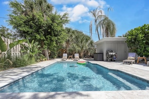 A rectangular swimming pool with clear blue water, surrounded by lush greenery and palm trees. There are two lounge chairs and a barbecue grill in the background. - Starfish Contemporary Home (Sarasota)
