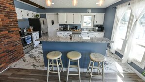 A kitchen with a central island, bar stools, white cabinets, and a brick wall. - Whispering Pines Manor

 (Johnstown)