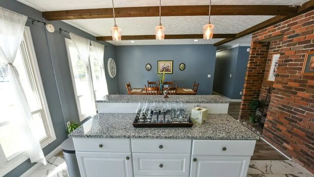 A kitchen with a granite countertop, white cabinets, a dining area with a table and chairs, and a brick accent wall. - Whispering Pines Manor

 (Johnstown)