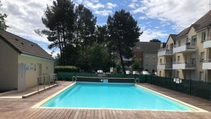A rectangular swimming pool with a wooden deck, surrounded by a green fence and trees. - Appartement en Résidence Sécurisée Circuit 2kms (Le Mans)