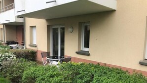 A building with a balcony, a small patio with a table and chairs, and a well-maintained hedge. - Appartement en Résidence Sécurisée Circuit 2kms (Le Mans)