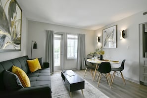 Ett modernt vardagsrum med en svart soffa, ett matbord med stolar och ett stort fönster med gardiner.