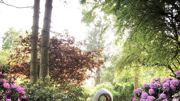 En have med en rund stenskulptur, omgivet af frodigt grønt og blomstrende buske.