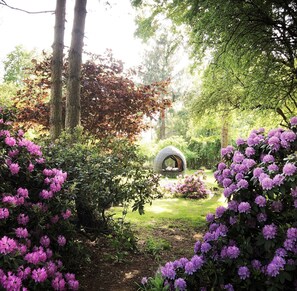 Un jardín con una estructura circular de piedra, rodeado de abundante follaje y arbustos florecientes.