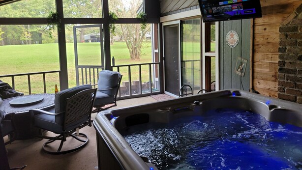 Ein Whirlpool mit an der Wand montiertem Fernseher, ein Terrassenstuhl und Blick auf einen grünen Garten durch große Fenster.