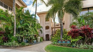 A hotel entrance with a palm tree-lined pathway leading to a building with a balcony. - Includes Beach Gear!  134 Shores at Waikoloa (Waikoloa Beach Resort)