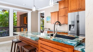 A modern kitchen with a marble countertop, wooden cabinets, stainless steel appliances, and a large window offering a view of greenery. - Includes Beach Gear!  134 Shores at Waikoloa (Waikoloa Beach Resort)