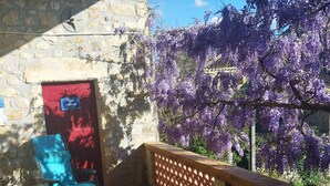 A covered outdoor area with a stone wall, a red door with a sign, a blue chair, and purple flowering trees. - mas Cévenol en Ardèche Animaux Acceptés (rosieres)