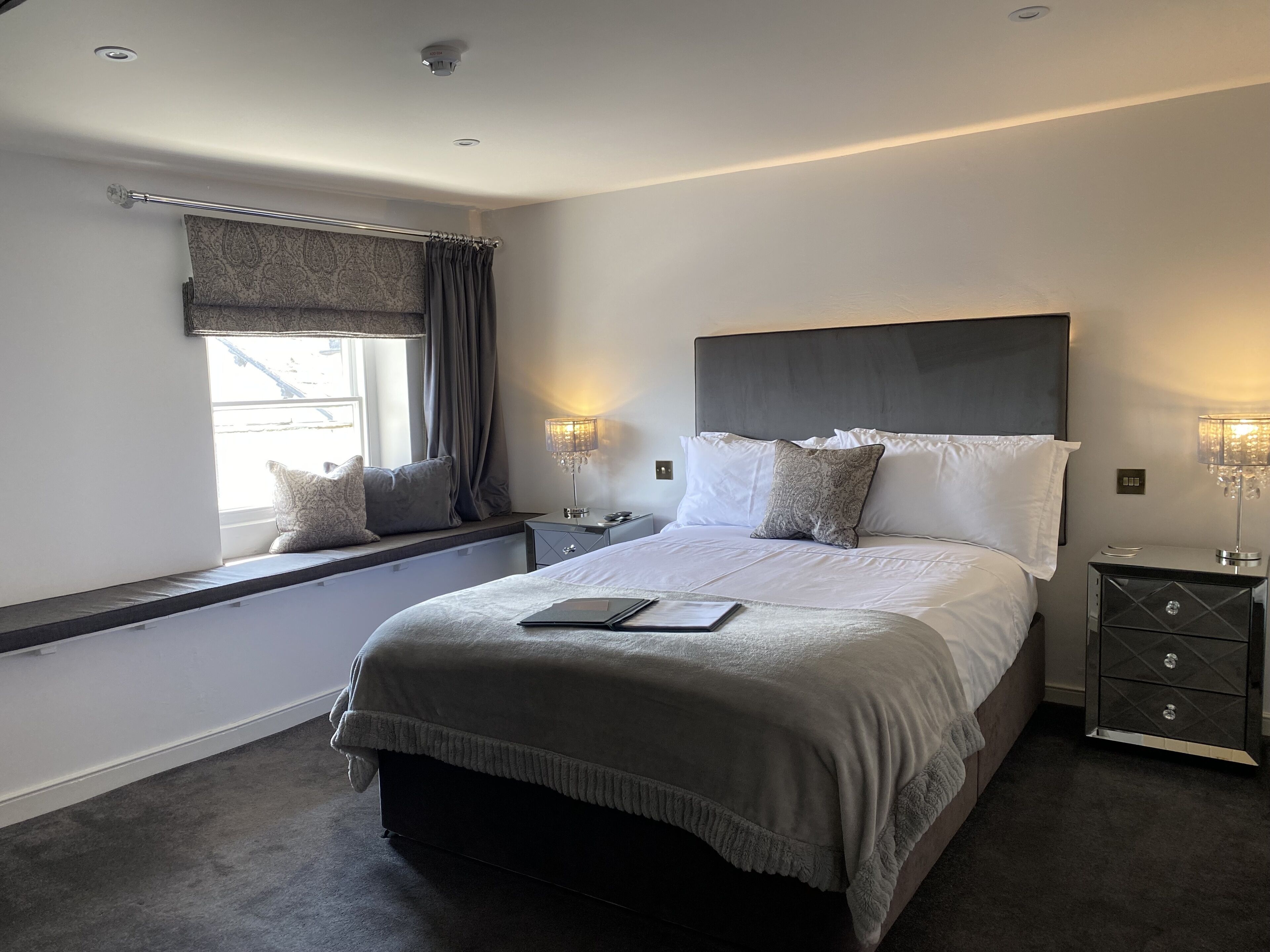 Quarto moderno com uma cama grande, luminárias de cabeceira, uma janela com cortinas e uma mesinha de cabeceira com gavetas.