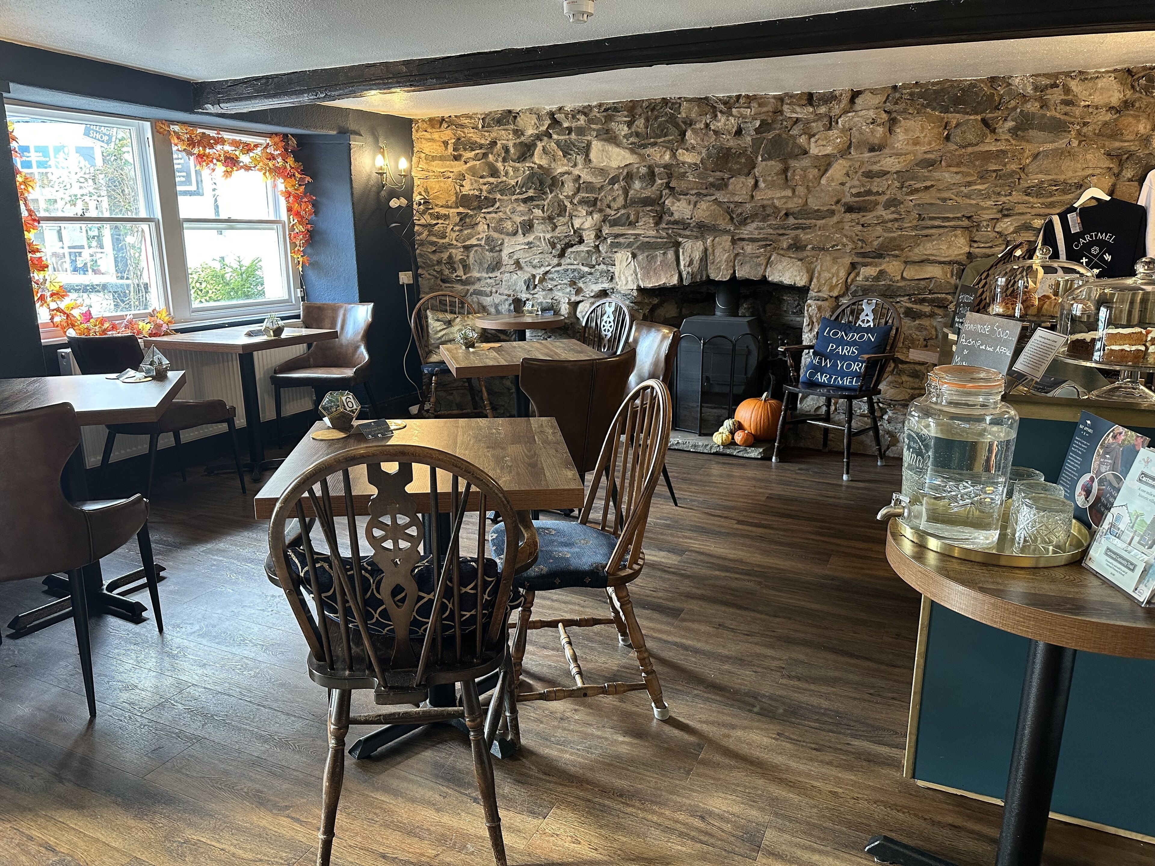 A cozy restaurant with wooden furniture, a stone wall, and a fireplace. Tables and chairs are arranged for dining.