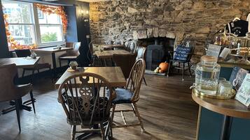 A cozy restaurant with wooden furniture, a stone wall, and a fireplace. Tables and chairs are arranged for dining.