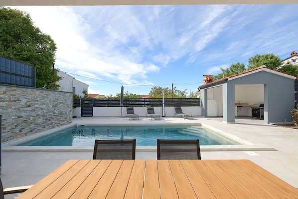 Relax by the pool surrounded by a low wall and a black fence, with a patio area offering a grill and kitchen nearby.