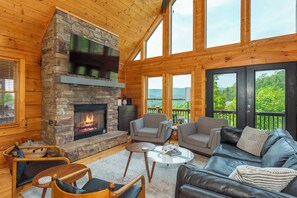 A log cabin interior with a stone fireplace, wooden walls, and a balcony with a view. - Big Views • Near Asheville • New Hot Tub with Mountain Views • New Weber Grill (Mars Hill)