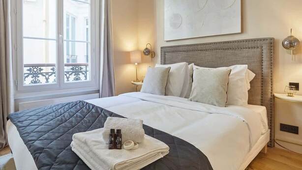 A hotel room with a bed, a nightstand, a lamp, and a window with a view of buildings. - Sublim new flat center of Paris Opéra Madeleine (Paris)