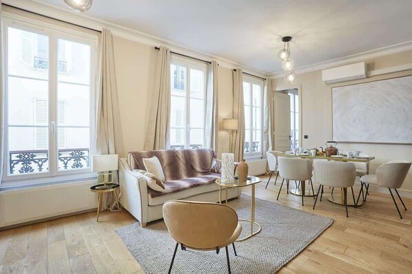 A modern living room with a sofa, coffee table, and chairs. There is a dining area with a table and chairs. Large windows allow natural light to enter. - Sublim new flat center of Paris Opéra Madeleine (Paris)