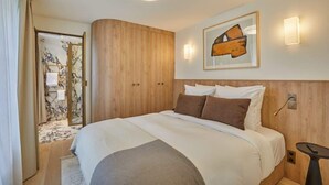 A hotel room with a bed, wooden wardrobe, a framed artwork, and a small table.