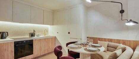 A modern kitchen with a dining area, featuring a built-in oven, a round dining table with a light fixture above, and a sofa with a wooden accent wall.