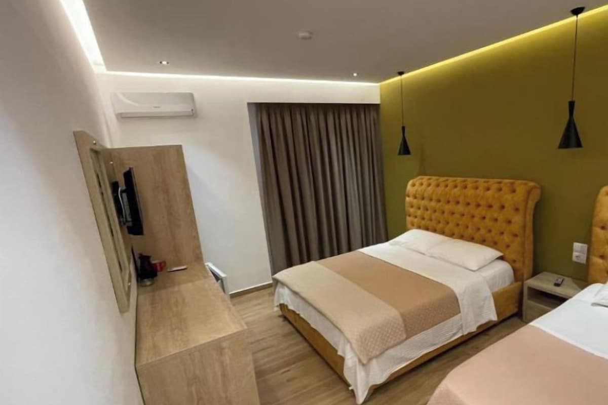 Ein modernes Hotelzimmer mit Holzboden, einem Bett mit beige-weißer Bettdecke, einem zweiten Bett mit rosa Bettdecke, einer Holzbank, einem Spiegel und einem Fenster mit Vorhängen.