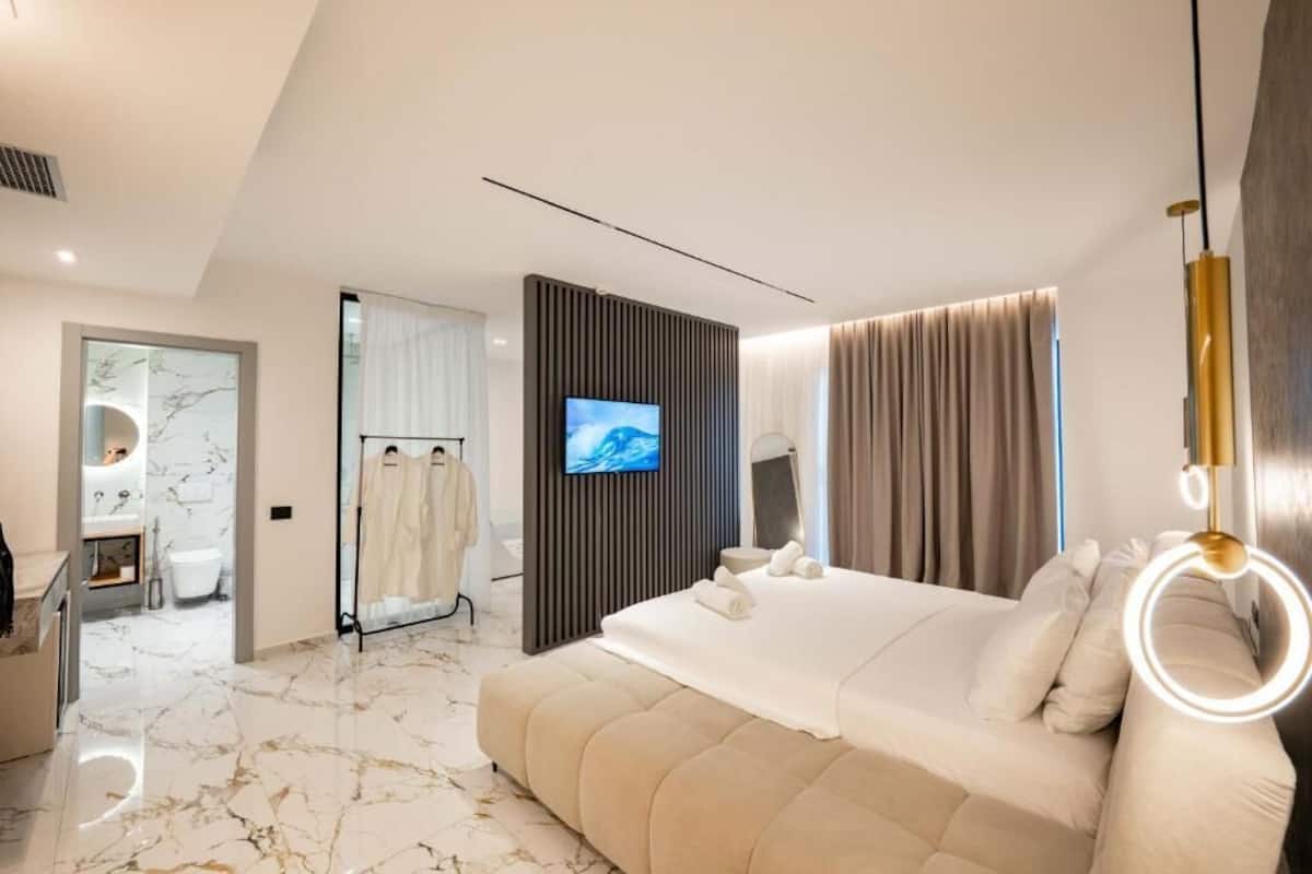 A modern hotel bedroom with a large bed, a TV, and a bathroom visible in the background.